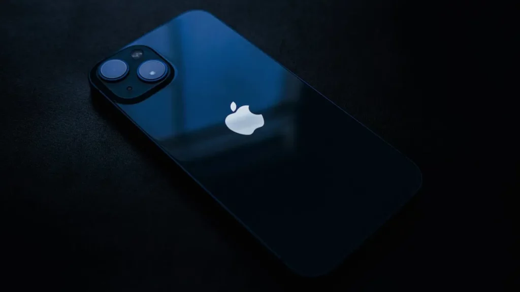 El iPhone 13, en color azul, en una mesa de madera, mostrando la pantalla de inicio con varias aplicaciones de iOS 18.